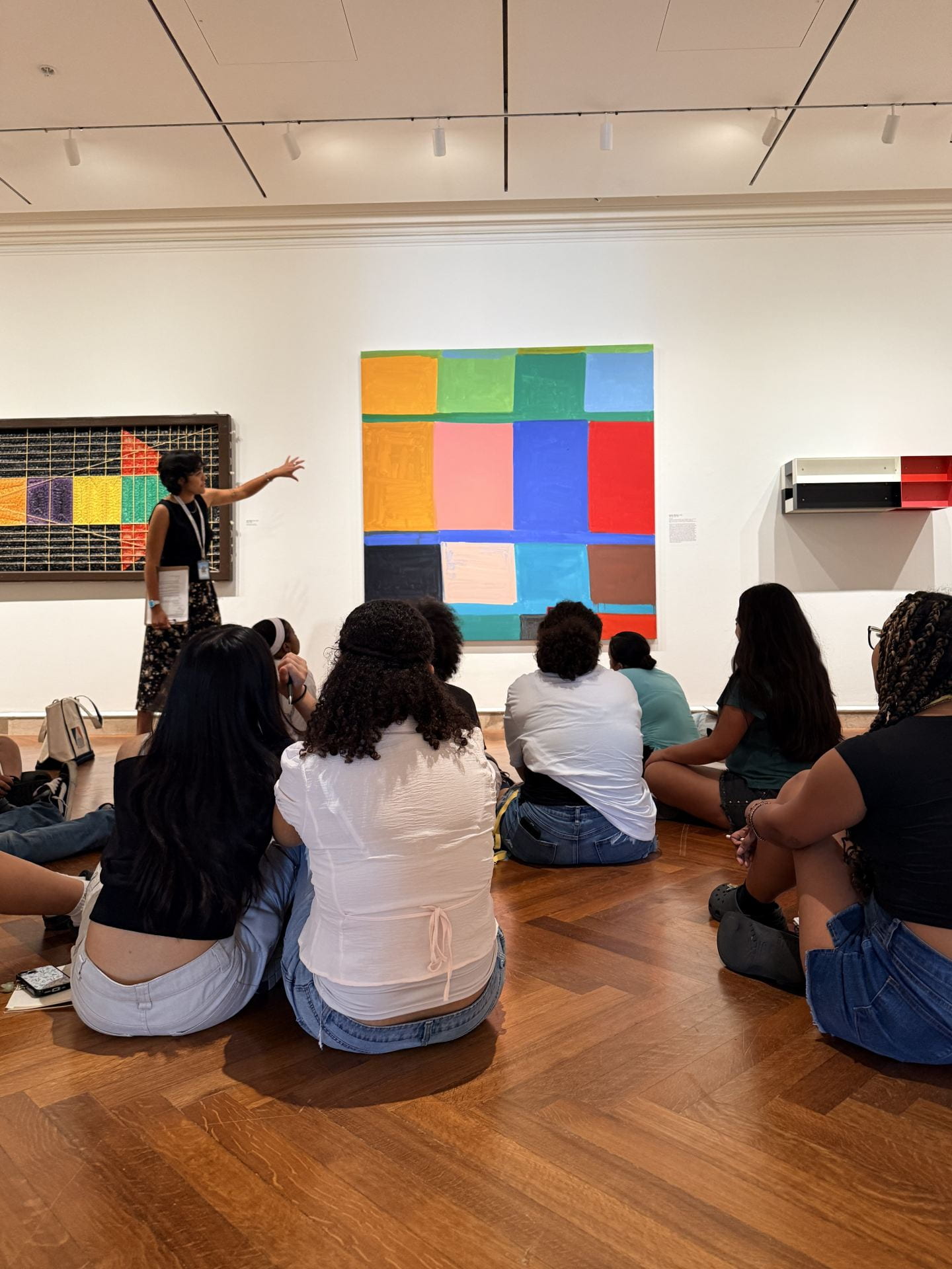 Museum Educator at top left, presenting works of art in a museum to adolescents who are seated on the floor of the gallery.